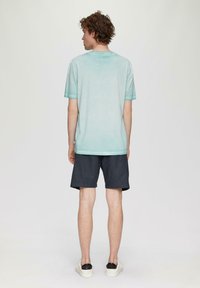 Gai zils īsām piedurknēm T-krekls ar smalku tekstūru, apvienots ar tumšiem šortiem. Modelis redzams no aizmugures, valkājot baltas slip-on kurpes.