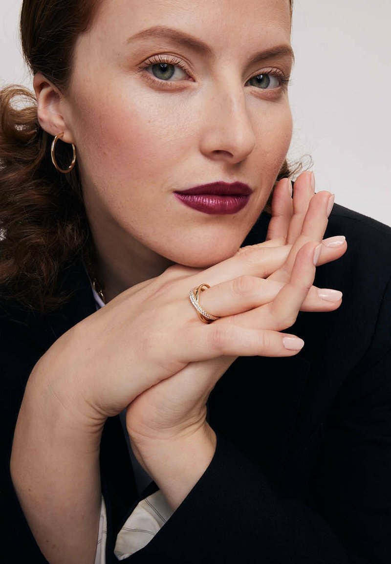 Modelka o jasnej karnacji, nosząca złote kolczyki w kształcie koła oraz skręconą złotą pierścionek. Ma na ustach ciemnoczerwoną pomadkę i czarną marynarkę na białej bluzce.