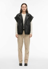 Veste sans manches en cuir noir avec col oversize, associée à une chemise à manches longues crème et un pantalon beige, posant sur un fond uni.