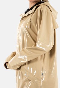 Impermeable beige brillante con capucha, botones de presión y un cuello interior negro. Presenta un diseño elegante y una textura suave.