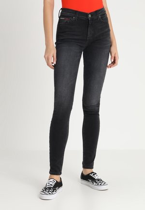 Kobieta ubrana w czarne obcisłe dżinsy, czerwoną bluzkę i czarno-białe trampki z wzorem płomieni, stojąca na tle białego tła.