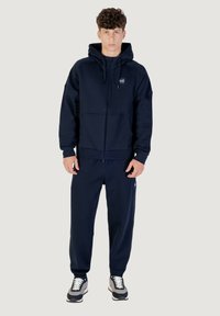 Dunkelblauer Zip-Hoodie mit Kängurutasche und passender Jogginghose. Hergestellt aus weichem Stoff, mit einem kleinen Logo auf der oberen linken Brust.