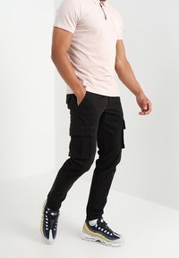 Czarne spodnie cargo z wieloma kieszeniami, zestawione z jasnoróżową koszulą polo. Obuwie w niebiesko-żółtym wzorze z siatkową teksturą.