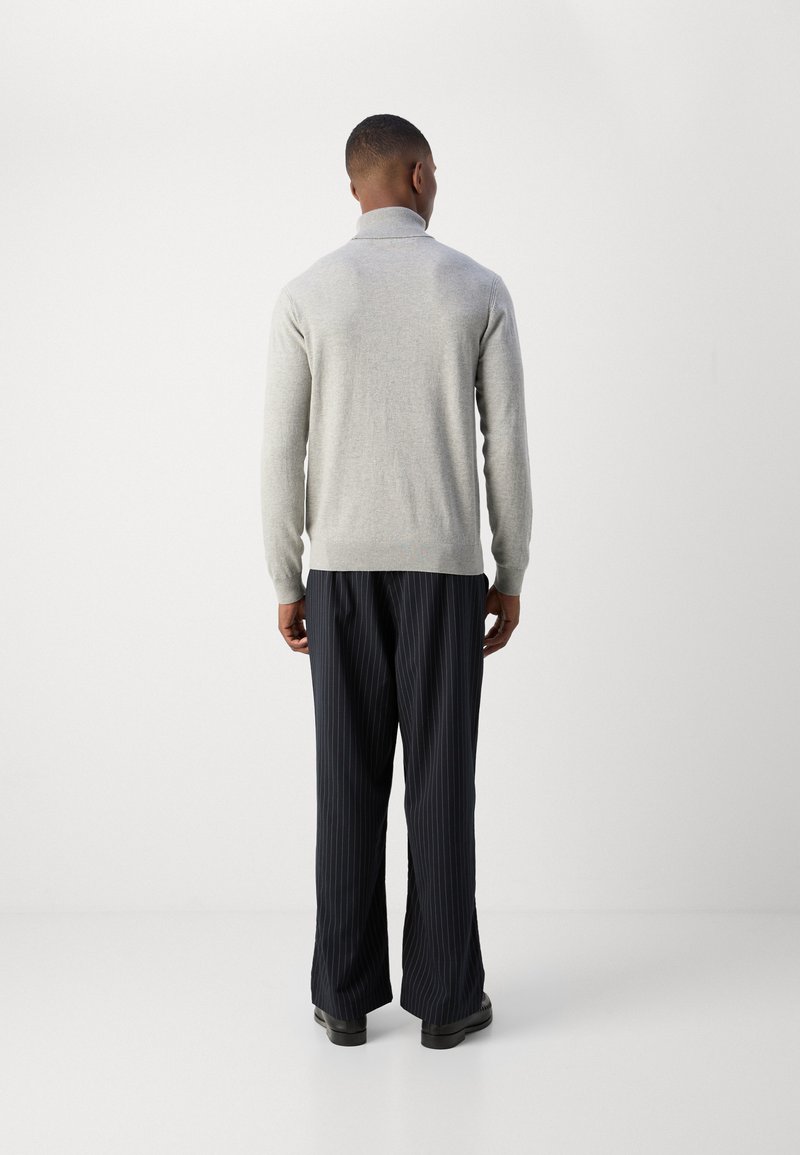 Pull en tricot gris clair avec un col montant, associé à un pantalon ample à rayures sombres et des chaussures noires. Vu de dos.