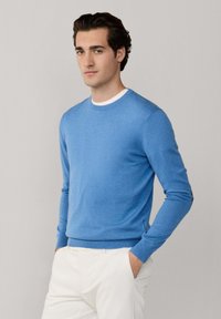 Młody mężczyzna stojący z rękami w kieszeniach, ubrany w jasnoniebieski sweter na białą koszulę i białe spodnie na jednolitym tle.