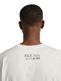 Mężczyzna o krótkich włosach i kolczykach-kołach, ubrany w białą koszulę z nadrukiem „F.U.T. Physical Education & Training B-583” na górnej części pleców.