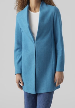 Cappotto in lana blu cielo con design senza colletto, maniche lunghe e taglio dritto, abbinato a una maglietta bianca e jeans di denim.