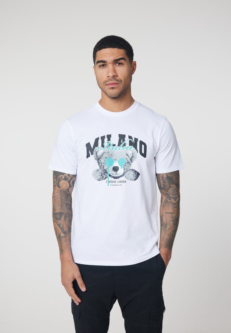 White cotton t-shirt featuring a bear graphic with text "MILANO Italia" in black and aqua colors. Short sleeves, round neckline, relaxed fit.