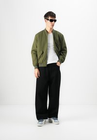 Oliwkowa kurtka bomber, biała koszulka, czarne szerokie spodnie i białe sneakersy z niebieskimi akcentami. Modelka w czarnych okularach przeciwsłonecznych.