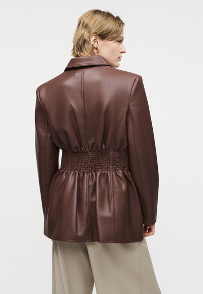 Άτομο με κοντά μαλλιά που φοράει καφέ δερμάτινο μπουφάν με ελαστική ζώνη στη μέση, γυρισμένο προς τα πίσω, μπροστά σε απλό λευκό φόντο.