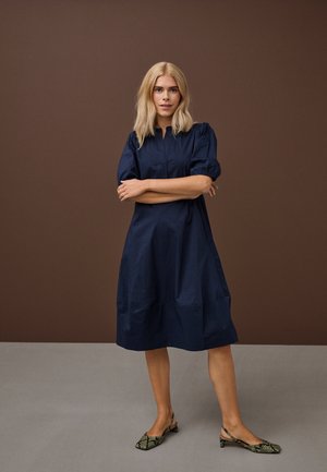 Blondinka z razpuščenimi lasmi stoji z rokami prekrižanimi, oblečena v mornarsko modro obleko do kolen in nizke pete z vzorcem kačje kože.