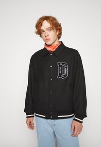 Black jacket featuring white striped cuffs and hem, with a prominent embroidered patch on the chest. Model wears a contrasting orange bandana.