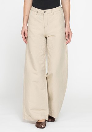 Donna che indossa pantaloni beige a gamba larga con chiusura con bottone e cerniera, tasche frontali e scarpe piatte intrecciate scure, in piedi su sfondo bianco.