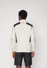 Langermet treningsskjorte i lys grå med svarte detaljer, med høy krage og en tettsittende design, kombinert med mørke shorts.