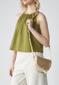 Top halter verde oliva con scollatura elastica, abbinato a pantaloni bianchi. Borsa a tracolla dorata con corpo in paglia testurizzata e patta piatta.