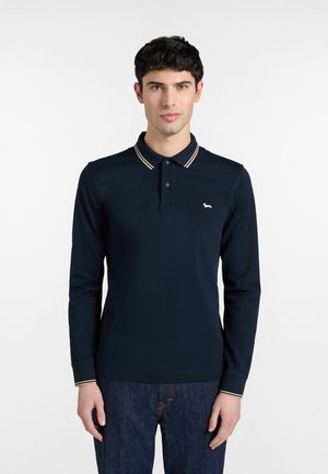 Mladić obučen u tamnoplavu polo majicu dugih rukava s prugastim ovratnikom i manšetama, uparenu s tamnoplavim trapericama, stoji na bijeloj pozadini.