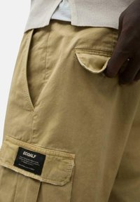 Pantalones cargo beige hechos de lona de algodón con un bolsillo frontal y una etiqueta que lleva "ECOALF". Presentan un corte relajado y costuras visibles.