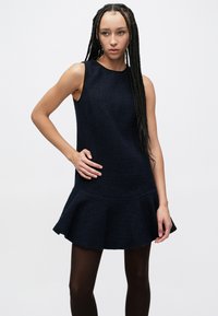 Temno modra obleka brez rokavov z teksturiranim materialom, razširjenim spodnjim delom in oprijetim zgornjim delom, združena s temnimi nogavicami.