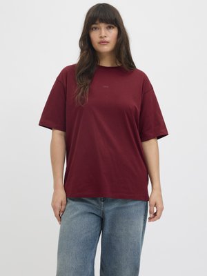 Femme aux longs cheveux foncés portant un t-shirt bordeaux ample et un jean bleu clair sur un fond clair uni.
