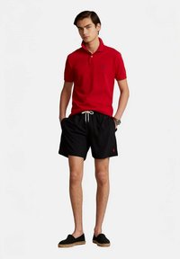 Ung mann iført en rød polo skjorte, svarte shorts med snor, og svarte slip-on espadrillos som står med hendene i lommene.
