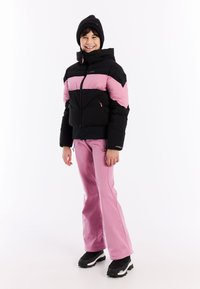 Zwart en roze geïsoleerde jas met een capuchon. Roze broek. Zwarte laarzen met grijze accenten. Glimlachend kindermodel staat tegen een witte achtergrond.