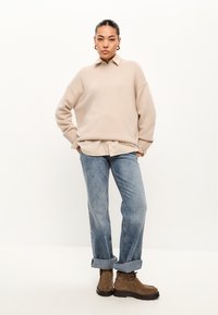 Beige gebreide trui met een kraag, een licht overhemd eronder, vervaagde blauwe jeans en bruine enkellaarsjes. Relaxed fit en opgerolde mouwen op de jeans.