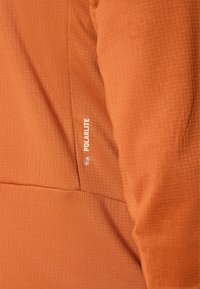 Oransje topp laget av teksturert stoff med en sydd logo "POLARLITE" i hvitt på siden. Designet har elegante linjer og minimal detaljering.