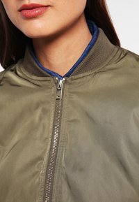 olivově zelená bomber bunda se zipem, vroubkovaným límcem a modrou podšívkou; hladká textura s mírně lesklým povrchem.