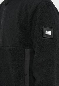 Black fleece jacket featuring a textured fabric, zippered collar, and a rectangular black label with white text on the sleeve.