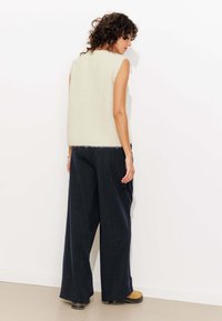 Haut en tricot sans manches ivoire avec une texture duveteuse, associé à un pantalon large à rayures bleu marine et des chaussures brunes. Arrière-plan neutre.