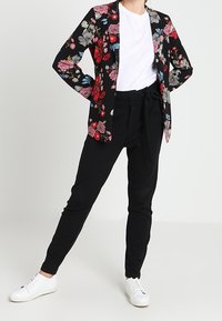 Černý květinový blejzr s růžovými a modrými květinami, kombinovaný s bílým tričkem a černými kalhotami. Model má na sobě bílé tenisky.