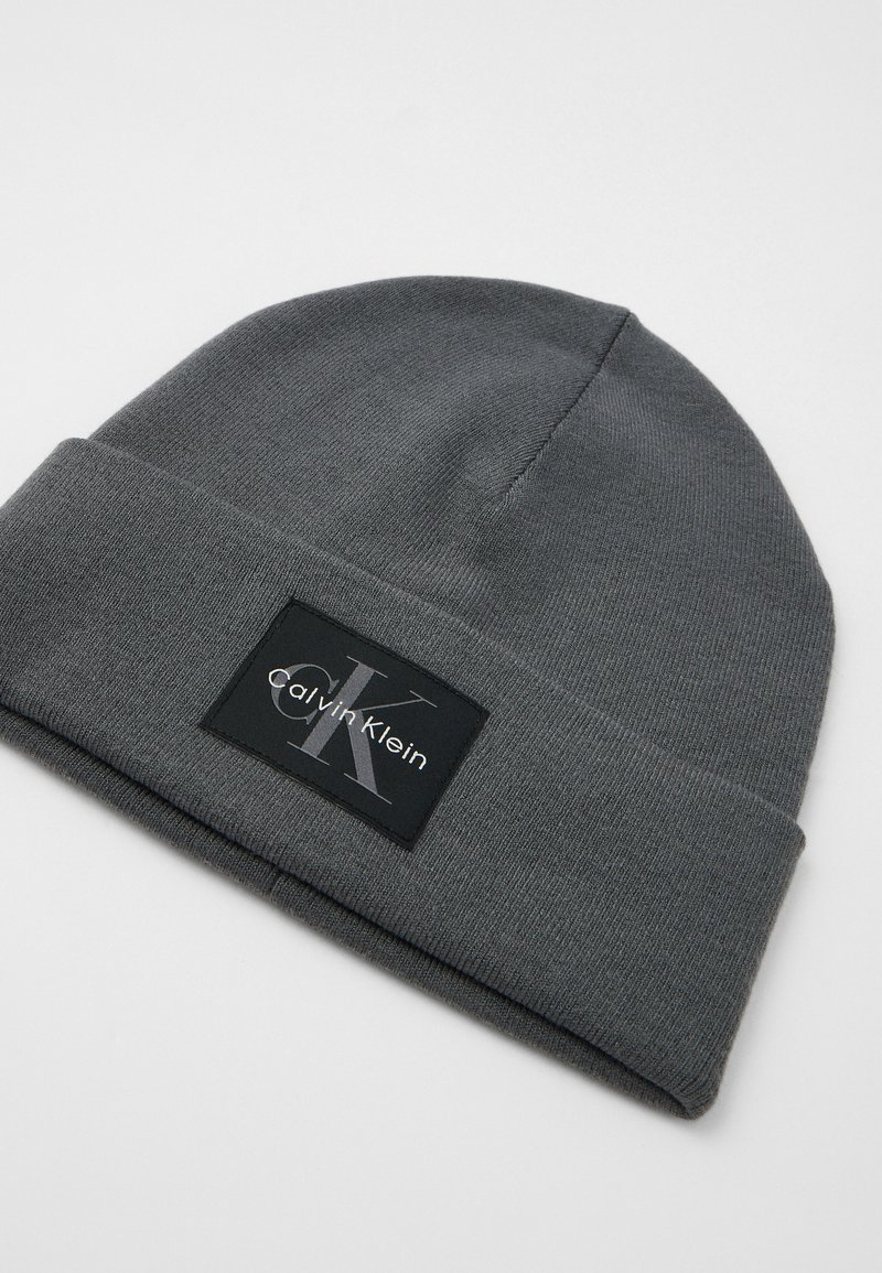 Bonnet en tricot gris avec un revers, arborant un patch rectangulaire noir avec le logo Calvin Klein en argent. Texture douce, design décontracté.