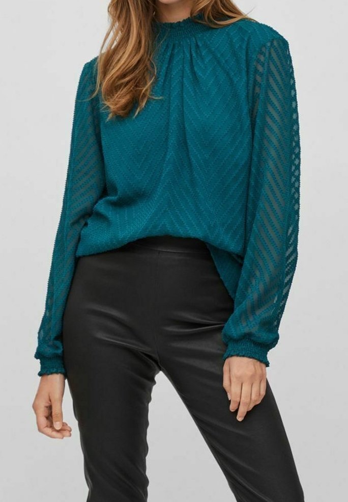 Blusa lunga teal a maniche lunghe in tessuto testurizzato con scollatura arricciata e maniche rigate trasparenti, abbinata a pantaloni neri aderenti.