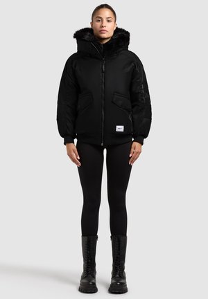 Giubbotto imbottito nero con cappuccio in pelliccia sintetica, chiusura con zip e due tasche frontali. Abbinato a leggins neri e stivali neri chunky.