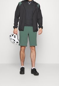 Černá lehká bunda s zelenými akcenty, černé tričko, zelené kraťasy a černé sportovní boty. Drží bílou helmu s ventilačními otvory.