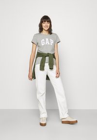 Pilkas marškinėliai su balta "GAP" logotipu, derinami su baltais džinsais ir žaliu megztiniu, surištu ant liemens. Šviesiai smėliniai batai su ruda neišorinėmis dalimis.