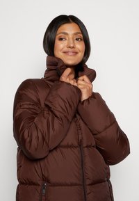 Braune Pufferjacke mit hohem Kragen, Reißverschlusstaschen, gestepptem Design und glatter, glänzender Textur. Verstellbarer Halsverschluss sichtbar.