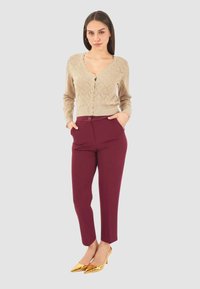 Donna in piedi con le mani in tasca, che indossa un cardigan beige con bottoni, pantaloni bordeaux e tacchi alti a punta dorati, su uno sfondo grigio.