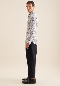 Weißes Blumen-Hemd mit Knopfschluss, das mit blauen, roten und grünen Mustern verziert ist, kombiniert mit schwarzen, knöchellangen Hosen und schwarzen Schuhen.