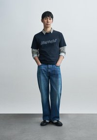 Marineblauw katoenen t-shirt met grafische tekst, gecombineerd met een geruit overhemd, wijd uitlopende blauwe spijkerbroek en zwarte schoenen.