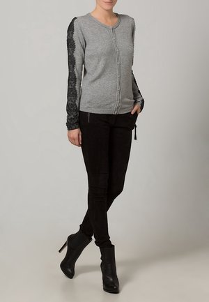 Cardigan gris avec détails en dentelle sur les manches, fermeture par boutons, col rond. Porté avec un pantalon noir ajusté et des bottines.