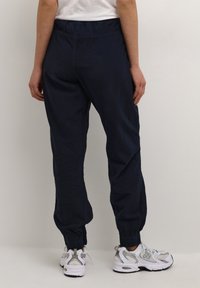 Marinblå joggers med elastisk midja och smalnande ärmar, tillverkade av lättviktsmaterial. Bärs med grå och vita träningsskor.