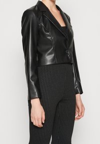 Leikattu musta keinonahkainen bleiseri lovitetulla kauluksella, yksinapaisella napituksella ja ryhdikkäillä hartioilla, yhdistetty raidallisiin mustiin housuihin.