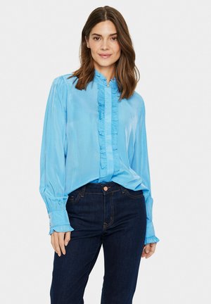 Blusa in satin blu con frontale arricciato, chiusura a bottoni e maniche lunghe con polsini arricciati. Abbinata a jeans di denim scuri.