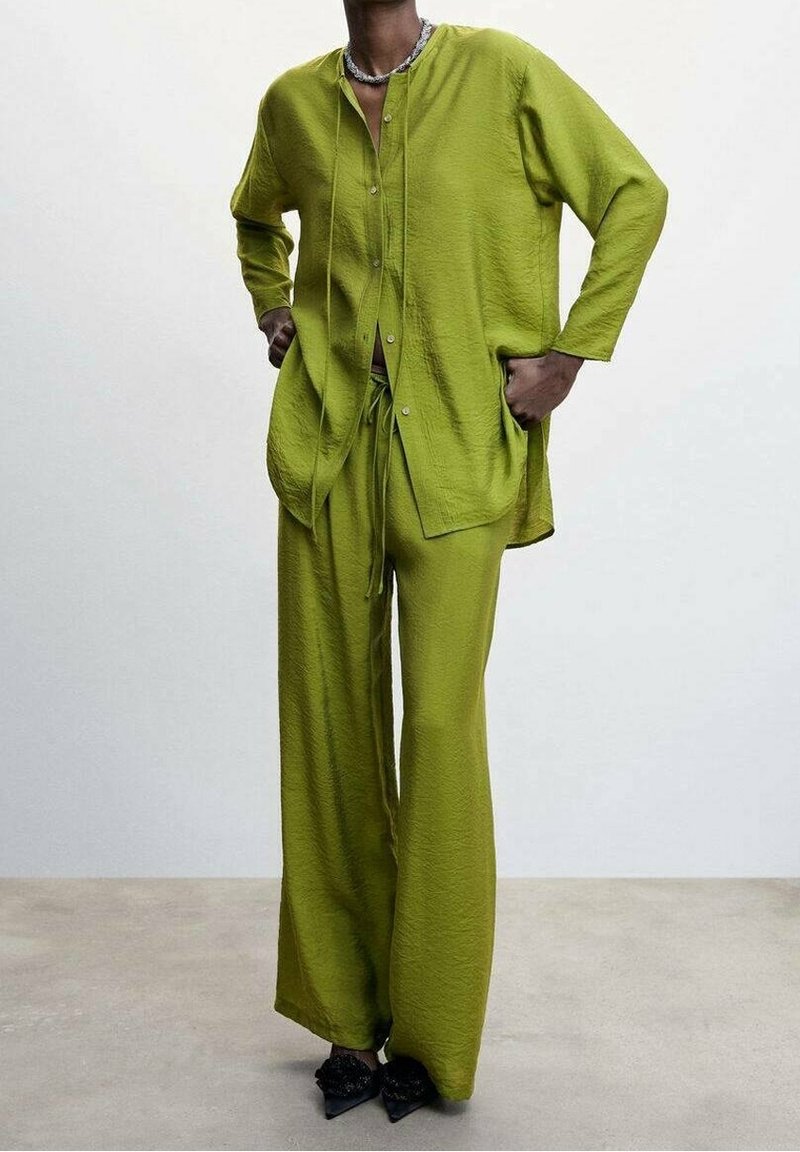 Personne portant une chemise ample texturée vert citron à boutons et un pantalon large assorti, debout les mains dans les poches devant un fond uni.