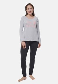 Donna in piedi a piedi nudi con una maglietta grigia a maniche lunghe del Pink Panther e leggings neri su uno sfondo bianco semplice.