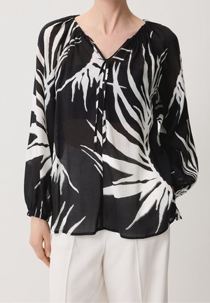 Blouse noire transparente avec des motifs abstraits de feuilles blanches, manches longues, encolure froncée avec un lien devant, portée avec un pantalon blanc.