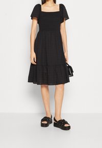 Schwarzes Kleid mit quadratischem Ausschnitt, kurzen Puffärmeln und gestuftem Rüschensaum. Aus strukturiertem Stoff gefertigt. Kombiniert mit schwarzen, klobigen Sandalen.