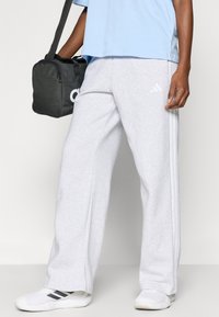 Szürke melegítőnadrág fehér csíkokkal, puha anyagból. Világoskék inggel és szürke táskával viselve. A nadrágon márkajelzés található.