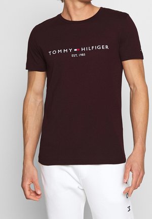 T-shirt en coton bordeaux avec col rond et manches courtes. Présente le logo blanc "TOMMY HILFIGER" et le texte "EST. 1985" sur la poitrine.